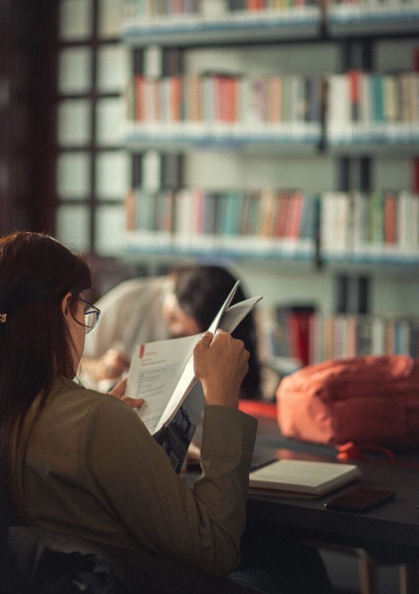 Adult woman reading in a cozy library setting, surrounded by bookshelves.